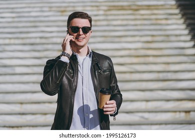 Young Man In Leather Jacket Eyeglasses Walk In City Downtown Go Down Concrete Steps Talk Speak By Mobile Cell Phone Takeaway Craft Paper Cup Drink Coffee To Go Look Aside Concept Of Urban Lifestyle