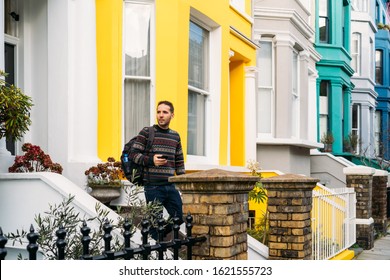 Young Man Leaving His House And Using His Smart Mobile With Colorful Houses On Portobello Street In The Notting Hill Neighborhood