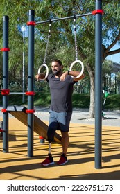 Young man with leg prothesis using gymnastic rings for training. Strong sportsman standing on sports ground holding rings with hands looking far. Health care, sports for people with disability concept