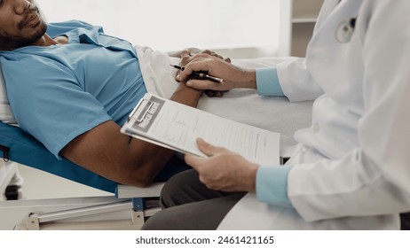 A young man lies on a bed in a hospital ward talking with her young Indian doctor. Doctor checking patient's health doctor treating patient, hospital admission of patient - Powered by Shutterstock