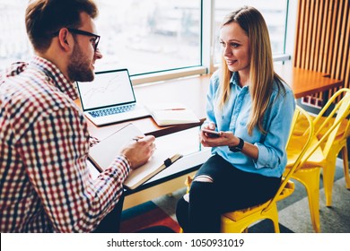 Young man listening female colleague with smartphone in hand and making some notes of creative ideas in notepad during consultancy.Creative student cunducting interview in stylish businesswoman