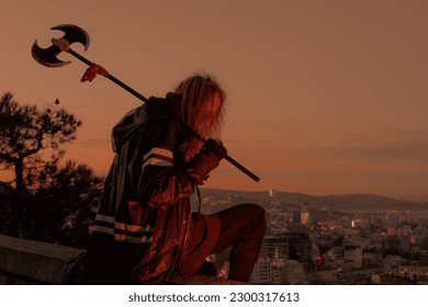 Young man long blond hair viking style with an ax in his hands, sitting above city on mountain top during sunset - Powered by Shutterstock