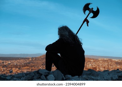 Young man long blond hair viking style with an ax in his hands, sitting above city on mountain top during sunset - Powered by Shutterstock