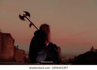 Young man long blond hair viking style with an ax in his hands, sitting above city on mountain top during sunset - Powered by Shutterstock