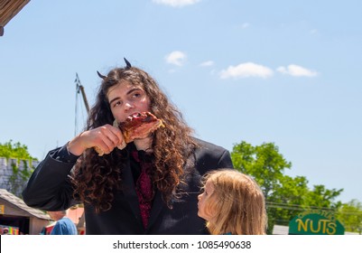 A Young Man With Long Curly Hair And Horns Bites Into A Turkey Leg While His Blond Child Looks On In Anticipation At The Oklahoma Renaissance Festival In Muskogee Oklahoma USA 5 5 2018