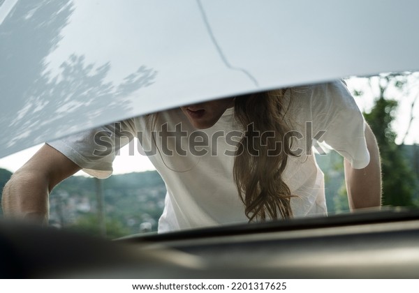 Young man with long hair looking inside\
under the hood of car, View from the inside of\
car