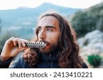 a young man with long hair plays the harmonica. a man enjoys playing the harmonica while standing on the street.