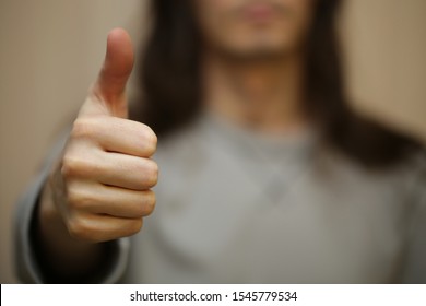 Young man with long hair shows his respect and approval by thumb up gesture. Serious guy without a smile on his face. Good job symbol. Shallow depth of field and  blurred background