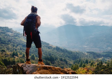 Young Man With Long Hair Without Shirt And Big Backpack On Rock In Mountain Looking The City. Man Standing In Shorts And Black Backpack. Man Bun.
