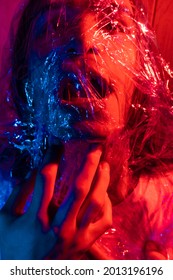  Young Man With Long Hair Wrapped In A Plastic Bag. Man Covered With Cellophane Wrap. Portrait In Blue And Red Lighting.