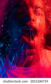  Young Man With Long Hair Wrapped In A Plastic Bag. Man Covered With Cellophane Wrap. Portrait In Blue And Red Lighting.