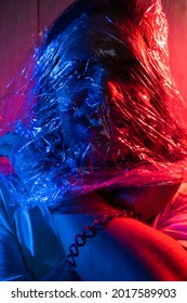  Young Man With Long Hair Wrapped In A Plastic Bag. Man Covered With Cellophane Wrap. Portrait In Blue And Red Lighting.