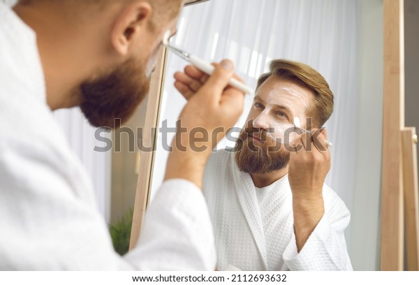 Young man look in mirror do facial skin procedures\
after shower at home. Male metrosexual apply cream or serum on\
face, make beauty anti-aging treatment. Skincare concept.\
Cosmetology and care.