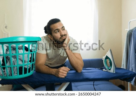 Young man looking bored while tackling housework, staring at a pile of clothes and contemplating the tedious task of ironing