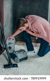 Young Man Looking At Broken Vacuum Cleaner At Home 