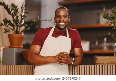 Young man looking at the camera with a bright smile while wearing a white apron in a cozy cafe. Male entrepreneur embodying success, hospitality, and small business owner vibes. - Powered by Shutterstock