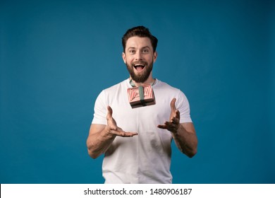 Young Man Looking Casual In Surprise Throwing Big Red Present Gift Box In Air On Blue Studio Background. Portrait Of A Handsome Man.