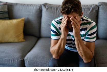 Young man looking depressed while sitting alone on his living room sofa at home with his face in hands