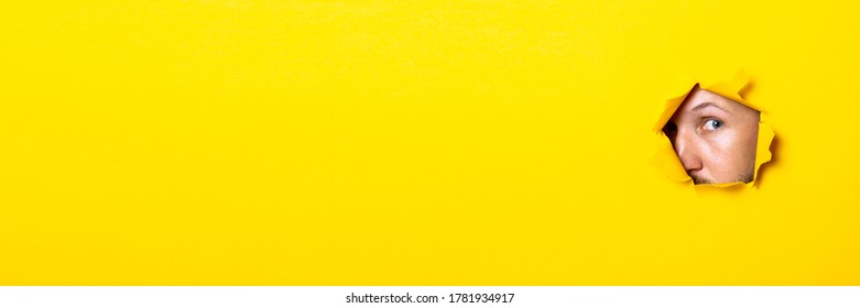 Young man looking with an eye into the hole of torn yellow background. Banner. - Shutterstock ID 1781934917