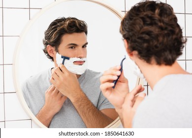 Young man looking at mirror while shaving on blurred foreground in bathroom