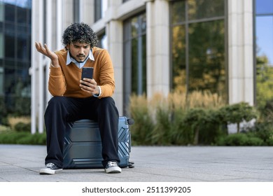 Young man looking at phone with confusion sitting on suitcase outside modern office building. Business professional appearing frustrated while waiting, balancing technology usage in urban setting. - Powered by Shutterstock