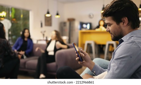 Young Man Looking At Smartphone Device In Resting Room At Workplace. Person Browsing Internet Social Media On Phone At Office Place