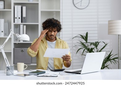 Young man looking stressed while reviewing documents in a modern home office setting with a laptop, coffee, and desk accessories.