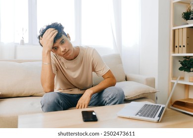 Young man looking stressed while sitting at home on a sofa, with a laptop and phone nearby, expressing frustration over work or personal issues in a bright, minimalistic setting - Powered by Shutterstock