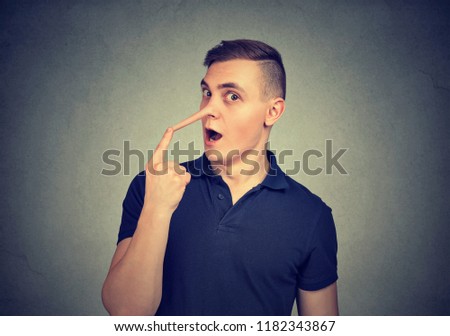 Young man looking surprised at camera touching long fake nose of liar on gray background