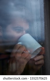 Young Man Looking Through A Window In A Rainy Day, Drinking Coffee At Home