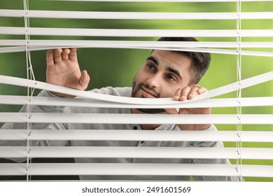 Young man looking through window blinds on blurred background