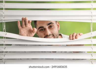Young man looking through window blinds on blurred background