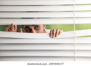 Young man looking through window blinds on blurred background