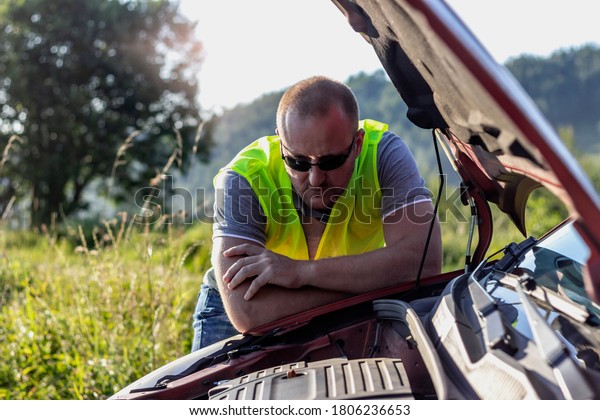 Young man looking under the hood of broken car on\
the road.Blond hair male standing by the broken car.Man having\
problem with his car.Broken car on the road.Man looking under the\
auto hood.Copy space.