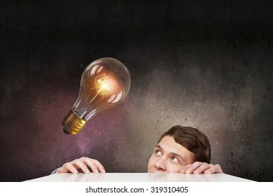 Young Man Looking From Under Table At Glass Light Bulb