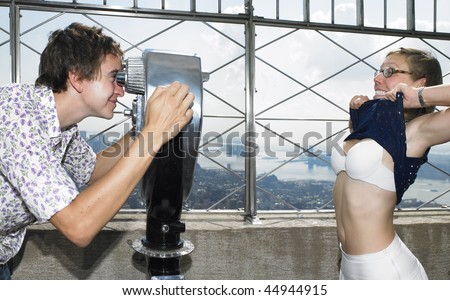 Young man looking at young woman through telescope as she lifts her top and shows her bra. Horizontal shot.