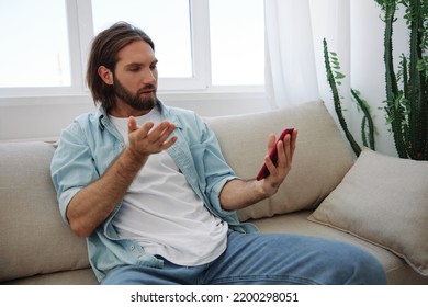 A Young Man Looks Angrily And Sadly At The Phone Screen At Home Sitting On The Couch. Bad News And Bad Luck At Work