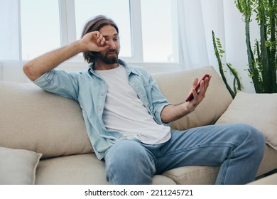 A Young Man Looks Angrily And Sadly At The Phone Screen At Home Sitting On The Couch. Bad News And Bad Luck At Work