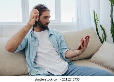 A Young Man Looks Angrily And Sadly At The Phone Screen At Home Sitting On The Couch. Bad News And Bad Luck At Work