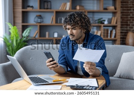 A young man looks concerned while reviewing a bill and using a mobile phone while sitting at his desk with a laptop at home.