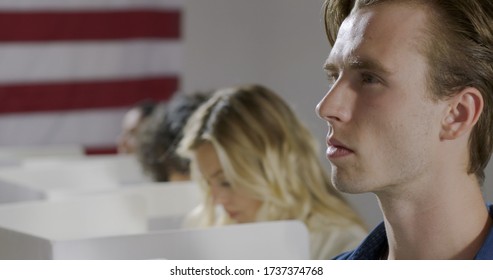 Young Man Looks Up To Think While Casting Vote With Others At Polling Station.  US Flag On Wall In Background.