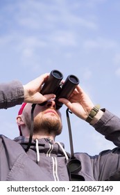 Young man looks up through binoculars against a blue sky, close-up. Birdwatching