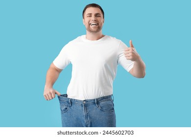 Young man in loose jeans showing thumb-up on blue background. Weight loss concept