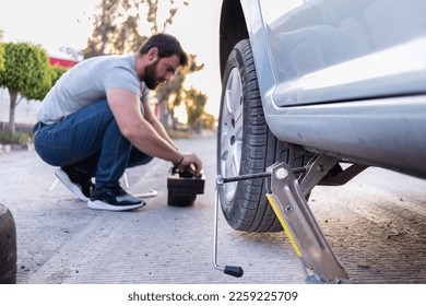 un joven aflojando una tuerca de su neumático de coche, cambiando un neumático plano en la carretera, se está usando un gato. joven aflojando una tuerca de su neumático de coche, cambiando una llanta en la carretera