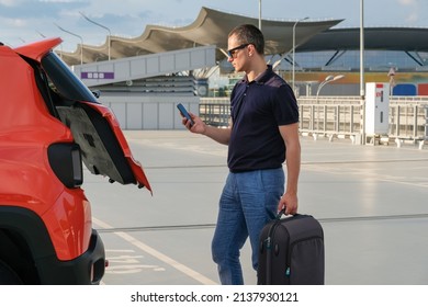 A Young Man With Luggage And A Phone In His Hand Stands Near His Car In The Parking Lot. Travel Concept.