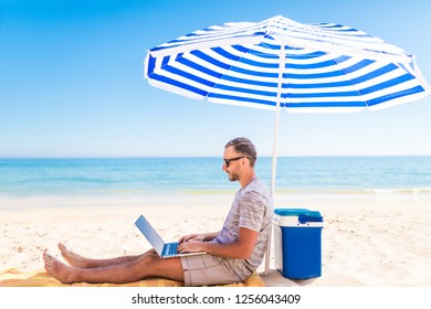 Young Man Lusing Laptop Computer On Beach Bench With Blue Umbrella Overhead, Surrounded By Turquoise Sea And Beautiful Sky.