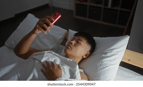 Young man lying in bed, using a smartphone, inside a bedroom at home, showcasing a relaxed and casual indoor setting in china, with a focus on modern lifestyle and technology