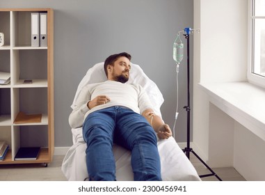 Young man lying in bed while receiving IV infusion in medical clinic. Male patient attached to intravenous drip getting vitamin treatment. Medical support and healthcare - Shutterstock ID 2300456941