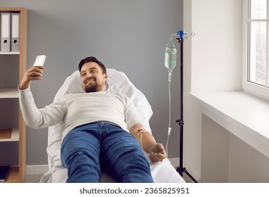 Young man lying in bed while receiving IV infusion and talking on phone. Male patient attached to intravenous drip getting treatment in medical clinic. Medical support and healthcare - Shutterstock ID 2365825951