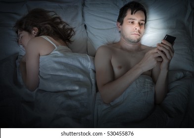 Young Man Is Lying With Girlfriend In Bed And Texting With Phone At Night. Insomnia And Cheating Concept.